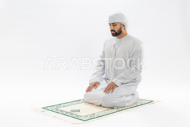 الخشوع أثناء أداء فريضة الصلاة، العبادة والتقرب إلى الله، أركان الإسلام الخمسة، بورتريه لرجل عربي خليجي إماراتي مسلم يرتدي الكندورة والعمامة يجلس على سجادة مخصصة، خلفية بيضاء