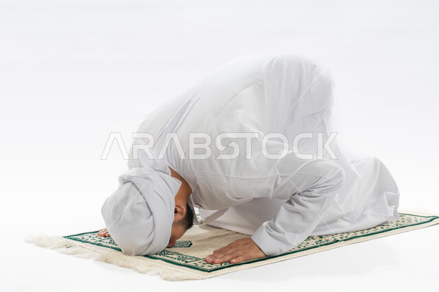 مفهوم العبادة والتقرب إلى الله، الخشوع أثناء أداء فريضة الصلاة، أركان الإسلام الخمسة، بورتريه لرجل عربي خليجي إماراتي مسلم يرتدي الكندورة والعمامة يجلس على سجادة مخصصة يقوم بالسجود، صورة لكامل الجسم، خلفية بيضاء