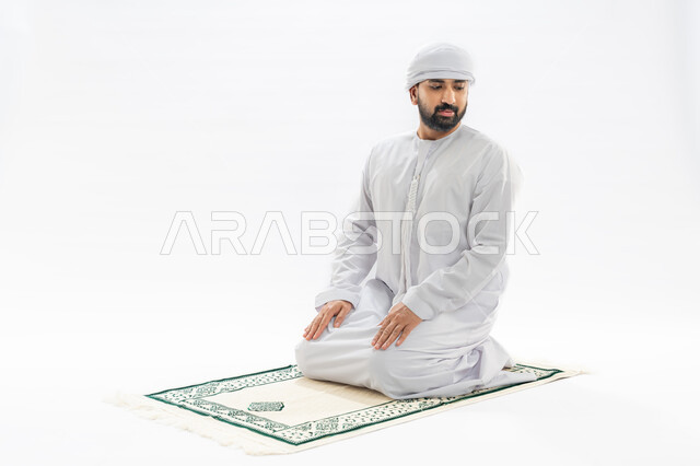 الخشوع أثناء أداء فريضة الصلاة، أركان الإسلام الخمسة، مفهوم العبادة والتقرب إلى الله، بورتريه لرجل عربي خليجي إماراتي مسلم يرتدي الكندورة والعمامة يجلس على سجادة مخصصة يقوم بأداء التحيات، صورة لكامل الجسم، خلفية بيضاء