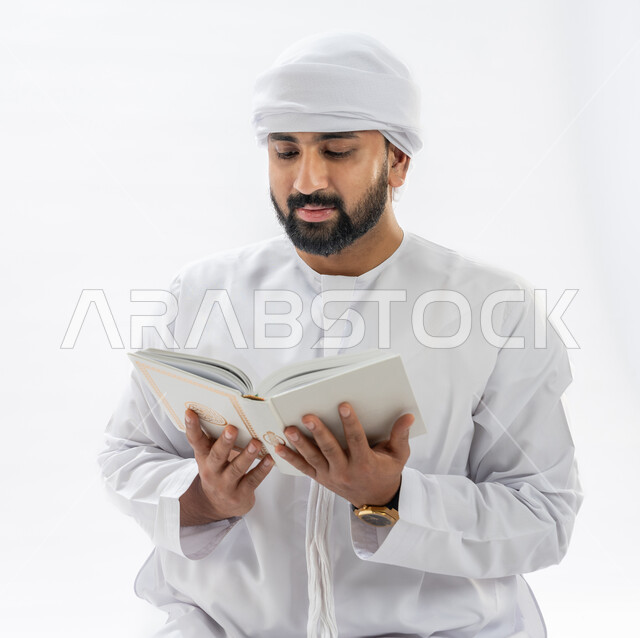 الخشوع أثناء تلاوة القرآن، مفهوم العبادة والتقرب إلى الله، بورتريه مقرب لرجل عربي خليجي إماراتي مسلم يرتدي الكندورة والعمامة يقوم بتدبر آيات المصحف الشريف، خلفية بيضاء