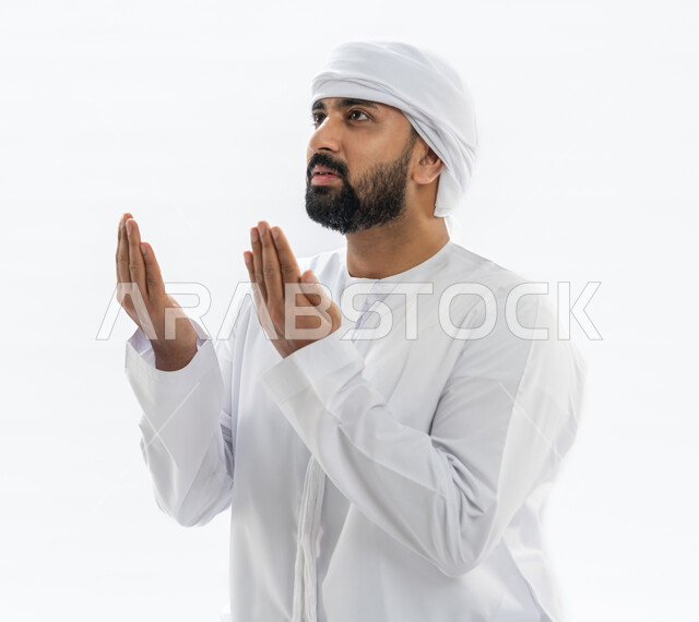إيماءات الخشوع بالدعاء، مفهوم العبادة والتقرب إلى الله، بورتريه لرجل عربي خليجي إماراتي مسلم يرتدي الكندورة والعمامة يقوم برفع يديه والتضرع إلى الخالق، أداء فريضة الصلاة، خلفية بيضاء