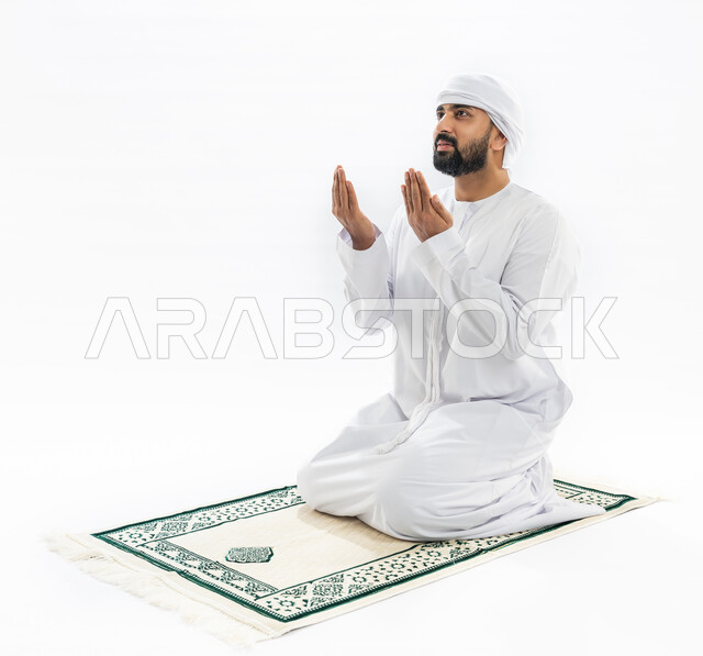 مفهوم العبادة والتقرب إلى الله، فع اليدين والتضرع إلى الخالق، إيماءات الخشوع بالدعاء، بورتريه لرجل عربي خليجي إماراتي مسلم يرتدي الكندورة والعمامة يصلي على سجادة مخصصة، أداء فريضة الصلاة، صورة لكامل الجسم، خلفية بيضاء