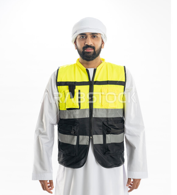 الاهتمام بالتطور العمراني بسواعد المهندسين الإماراتيين، مهن ووظائف هندسية، الوقوف باستقامة والنظر إلى الكاميرا بإيماءات البهجة والسرور, بورتريه لمهندس عربي خليجي إماراتي مبتسم يرتدي الكندورة والعمامة وسترة الحماية الخاصة بالعمل، خلفية بيضاء