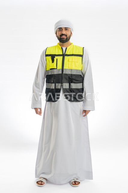 الوقوف باستقامة والنظر إلى الكاميرا بإيماءات البهجة والسرور, الاهتمام بالتطور العمراني بسواعد المهندسين الإماراتيين، بورتريه لمهندس عربي خليجي إماراتي مبتسم يرتدي الكندورة والعمامة وسترة الحماية الخاصة بالعمل، صورة لكامل الجسم، خلفية بيضاء