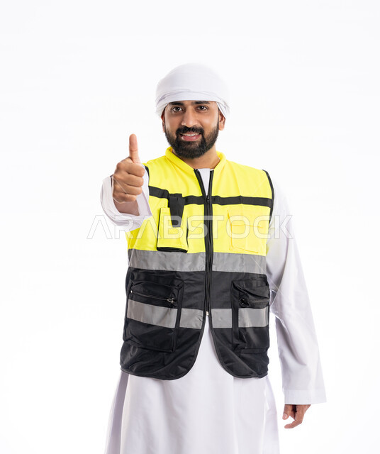 رفع اليد بإيماءات الجودة والامتياز، الوقوف باستقامة والنظر إلى الكاميرا بتعابير الرضا والقبول, الاهتمام بالتطور العمراني بسواعد المهندسين الإماراتيين، بورتريه لمهندس عربي خليجي إماراتي يرتدي الكندورة والعمامة وسترة الحماية، مهن ووظائف هندسية، خلفية بيضاء