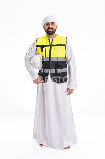 الوقوف باستقامة والنظر إلى الكاميرا بإيماءات البهجة والسرور, مهن ووظائف هندسية، الاهتمام بالتطور العمراني بسواعد المهندسين الإماراتيين، بورتريه لمهندس عربي خليجي إماراتي يرتدي الكندورة والعمامة وسترة الحماية  يمسك بيده الخوذة، صورة لكامل الجسم، خلفية بيضاء