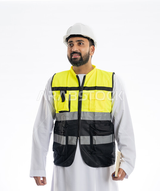 Looking at something with gestures of surprise, supervising work and following up on its progress, engineering professions and jobs, interest in urban development by the hands of engineers in the United Arab Emirates, portrait of an Arab Gulf Emirati engineer wearing a work jacket and a protective helmet holding a notebook, white background