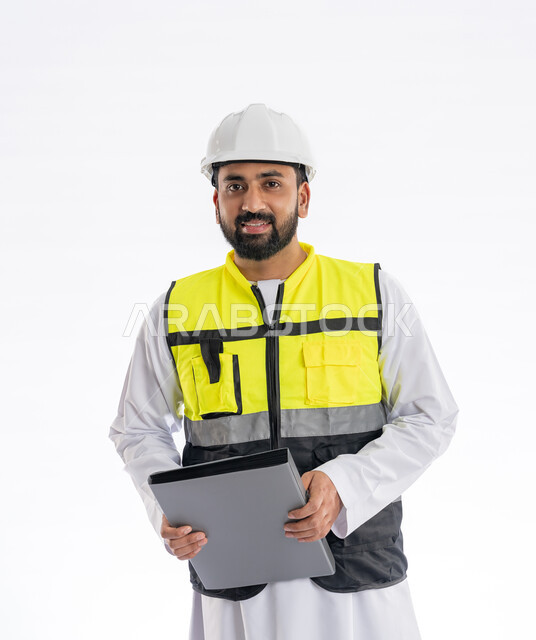 Engineering professions and jobs, following up on the progress of work and supervising it, looking at the camera with gestures of joy and pleasure, interest in urban development by the hands of engineers in the United Arab Emirates, portrait of an Arab Gulf Emirati engineer wearing a work jacket and a protective helmet holding construction records, white background