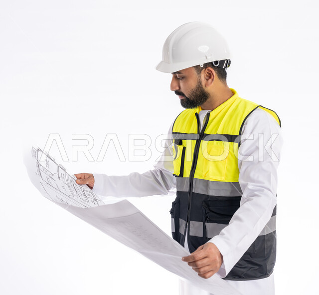 الإشراف على العمل ومتابعته، تدقيق مخططات المشروع، مهن ووظائف هندسية في الإمارات العربية المتحدة، بورتريه مقرب لمهندس عربي خليجي اماراتي يرتدي الكندورة وسترة وخوذة الحماية الخاصة بالعمل ينظر إلى مخطط هندسي بإيماءات التركيز، خلفية بيضاء