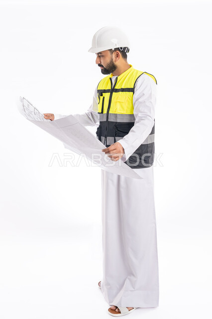 النظر إلى المخطط الهندسي بإيماءات التركيز، متابعة العمل والإشراف عليه، تدقيق مخططات المشروع، مهن ووظائف هندسية في الإمارات العربية المتحدة، بورتريه مقرب لمهندس عربي خليجي اماراتي يرتدي الكندورة وسترة وخوذة الحماية الخاصة بالعمل، صورة لكامل الجسم، خلفية بيضاء