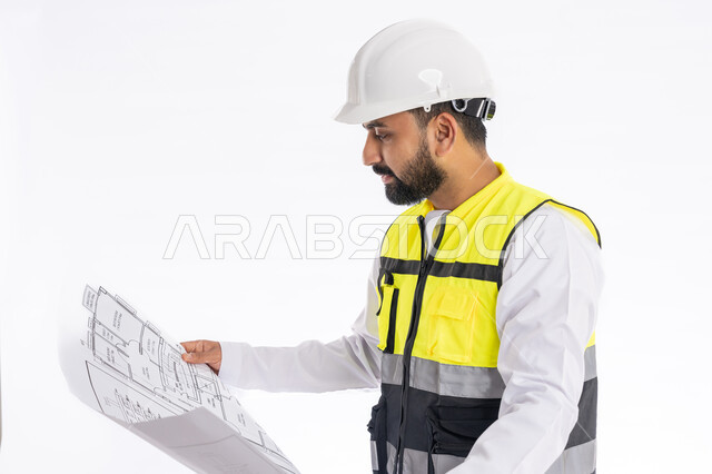 متابعة العمل والإشراف عليه، تدقيق مخططات المشروع، مهن ووظائف هندسية في الإمارات العربية المتحدة، بورتريه لمهندس عربي خليجي اماراتي يرتدي الكندورة وسترة وخوذة الحماية الخاصة بالعمل ينظر إلى مخطط هندسي بإيماءات التركيز، خلفية بيضاء