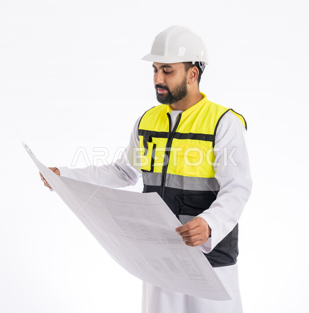 تدقيق مخططات المشروع، متابعة العمل والإشراف عليه، مهن ووظائف هندسية في الإمارات العربية المتحدة، بورتريه لمهندس عربي خليجي اماراتي يرتدي الكندورة وسترة وخوذة الحماية الخاصة بالعمل ينظر إلى مخطط هندسي بإيماءات التركيز، خلفية بيضاء