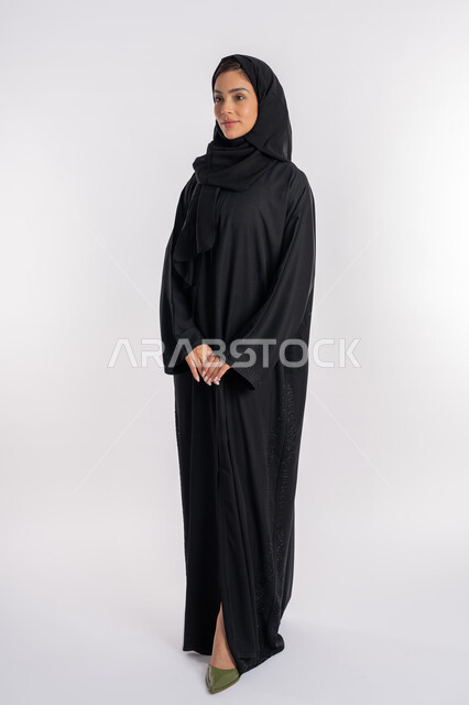 Young Saudi woman standing with clasped hands, modern elegant hijab fashion, concept of elegance and interest in outward appearance, portrait of a veiled Arab Gulf Emirati woman wearing a modern abaya and looking at something with gestures of pleasure and self-confidence, full body image, white background