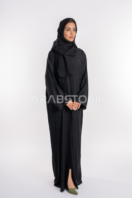 شابة سعودية تقف بأيدي متشابكة، أزياء محجبات عصرية راقية، مفهوم الأناقة والاهتمام بالمظهر الخارجي، بورتريه لامرأة عربية خليجية إماراتية محجبة ترتدي العباءة الحديثة وتنظر الى شيء ما بإيماءات السرور و الثقة بالنفس، صورة الجسم بالكامل، خلفية بيضاء