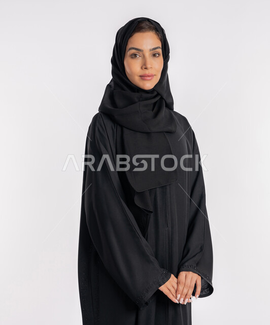 Modern and elegant veiled fashion, the concept of elegance and interest in external appearance, a close-up portrait of a veiled Arab Gulf Emirati woman wearing a modern abaya and looking at the camera with gestures of pleasure and self-confidence, a young Saudi woman standing with intertwined hands, white background