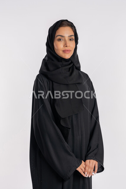 Modern and elegant veiled fashion, the concept of elegance and interest in external appearance, a close-up portrait of a veiled Arab Gulf Emirati woman wearing a modern abaya and looking at the camera with gestures of pleasure and self-confidence, a young Saudi woman standing with intertwined hands, white background