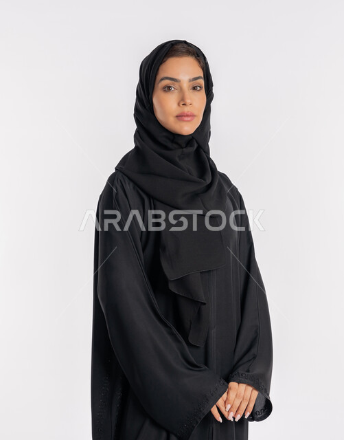 نظرات وتعابير الحزم والجدية، أزياء محجبات عصرية راقية، مفهوم الأناقة والاهتمام بالمظهر الخارجي، بورتريه مقرب لامرأة عربية خليجية إماراتية محجبة ترتدي العباءة الحديثة وتنظر الى الكاميرا بإيماءات الثقة بالنفس، شابة سعودية تقف بأيدي متشابكة، خلفية بيضاء
