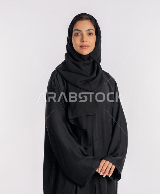 نظرات وتعابير السرور، أزياء محجبات عصرية راقية، مفهوم الأناقة والاهتمام بالمظهر الخارجي، بورتريه مقرب لامرأة عربية خليجية إماراتية مبتسمة ترتدي الحجاب والعباءة الحديثة وتنظر الى الكاميرا بإيماءات الثقة بالنفس، شابة سعودية تقف بأيدي متشابكة، خلفية بيضاء