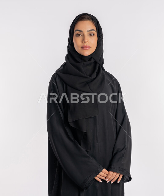 نظرات وتعابير الحزم والجدية، أزياء محجبات عصرية راقية، مفهوم الأناقة والاهتمام بالمظهر الخارجي، بورتريه مقرب لامرأة عربية خليجية إماراتية محجبة ترتدي العباءة الحديثة وتنظر الى الكاميرا بإيماءات الثقة بالنفس، شابة سعودية تقف بأيدي متشابكة، خلفية بيضاء