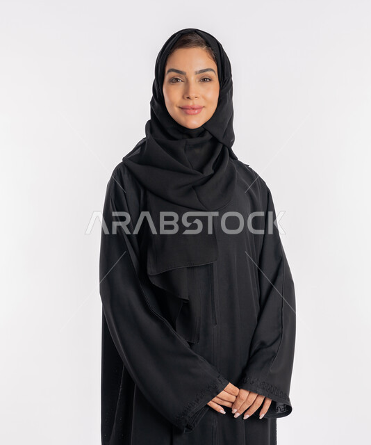 نظرات وتعابير السرور، أزياء محجبات عصرية راقية، مفهوم الأناقة والاهتمام بالمظهر الخارجي، بورتريه مقرب لامرأة عربية خليجية إماراتية مبتسمة ترتدي الحجاب والعباءة الحديثة وتنظر الى الكاميرا بإيماءات الثقة بالنفس، شابة سعودية تقف بأيدي متشابكة، خلفية بيضاء