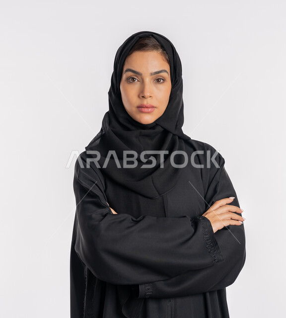 إيماءات الثقة بالنفس، أزياء محجبات عصرية راقية، مفهوم الأناقة والاهتمام بالمظهر الخارجي، بورتريه مقرب لامرأة عربية خليجية إماراتية محجبة ترتدي العباءة الحديثة وتنظر الى الكاميرا بتعابير الحزم والجدية، شابة سعودية تقف بأيدي متقاطعة، خلفية بيضاء
