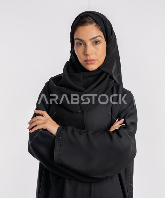 إيماءات الثقة بالنفس، أزياء محجبات عصرية راقية، مفهوم الأناقة والاهتمام بالمظهر الخارجي، بورتريه مقرب لامرأة عربية خليجية إماراتية محجبة ترتدي العباءة الحديثة وتنظر الى الكاميرا بتعابير الحزم والجدية، شابة سعودية تقف بأيدي متقاطعة، خلفية بيضاء