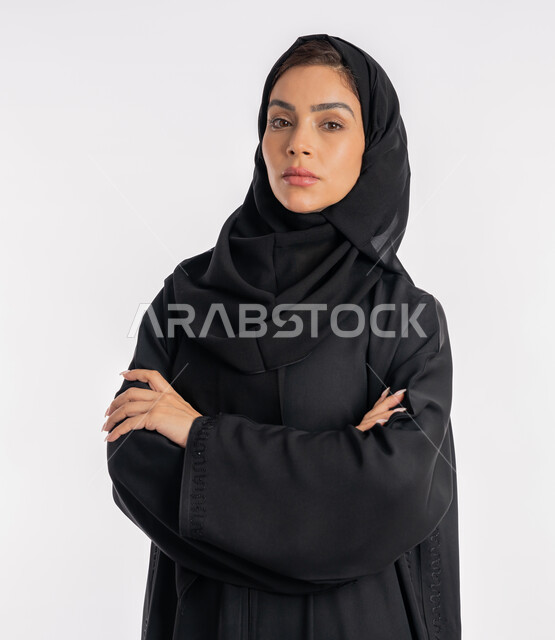 إيماءات الثقة بالنفس، أزياء محجبات عصرية راقية، مفهوم الأناقة والاهتمام بالمظهر الخارجي، بورتريه مقرب لامرأة عربية خليجية إماراتية محجبة ترتدي العباءة الحديثة وتنظر الى الكاميرا بتعابير الحزم والجدية، شابة سعودية تقف بأيدي متقاطعة، خلفية بيضاء