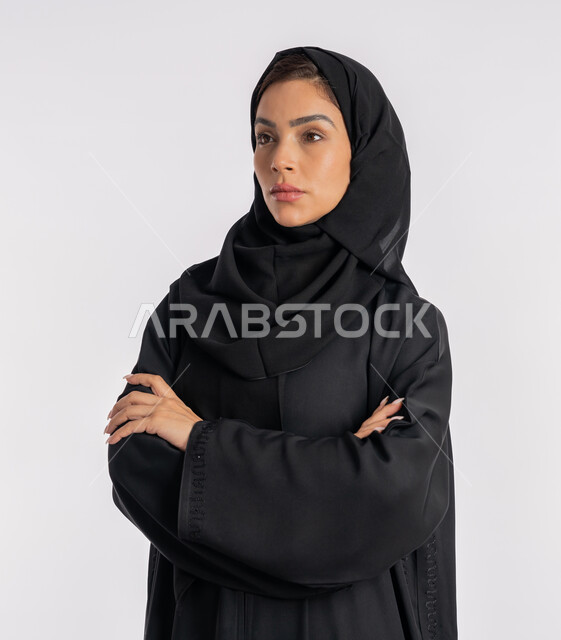 إيماءات الثقة بالنفس، أزياء محجبات عصرية راقية، مفهوم الأناقة والاهتمام بالمظهر الخارجي، بورتريه مقرب لامرأة عربية خليجية إماراتية محجبة ترتدي العباءة الحديثة وتنظر الى شيء ما بتعابير الحزم والجدية، شابة سعودية تقف بأيدي متقاطعة، خلفية بيضاء