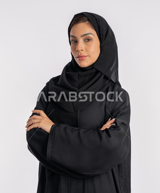 إيماءات الثقة بالنفس، أزياء محجبات عصرية راقية، مفهوم الأناقة والاهتمام بالمظهر الخارجي، بورتريه مقرب لامرأة عربية خليجية إماراتية محجبة ترتدي العباءة الحديثة وتنظر الى الكاميرا بتعابير الحزم والجدية، شابة سعودية تقف بأيدي متقاطعة، خلفية بيضاء