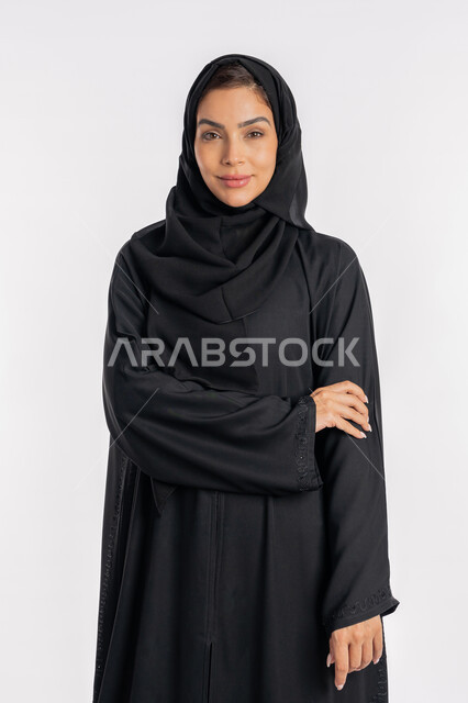 شابة سعودية تضع يدها اليمنى على ذراعها اليسرى، نظرات وتعابير السرور، أزياء محجبات عصرية ، مفهوم الأناقة والاهتمام بالمظهر الخارجي، بورتريه مقرب لامرأة عربية خليجية إماراتية مبتسمة ترتدي الحجاب والعباءة وتنظر الى الكاميرا بإيماءات الثقة بالنفس، خلفية بيضاء