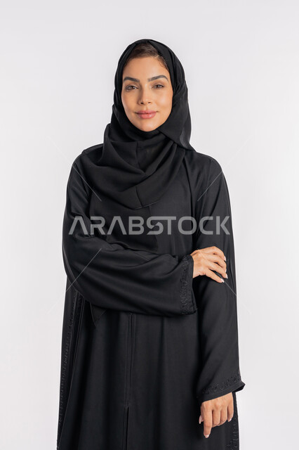 Young Saudi woman putting her right hand on her left arm, looks and expressions of pleasure, modern veiled fashion, concept of elegance and interest in outward appearance, close-up portrait of a smiling Emirati Gulf Arab woman wearing hijab and abaya and looking at the camera with gestures of self-confidence, white background