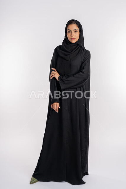 Saudi woman putting her left hand on her right arm, gestures of self-confidence, modern veiled fashion, elegance and interest in outward appearance, portrait of a young veiled Arab Gulf Emirati woman wearing a modern abaya and looking at the camera with expressions of firmness and seriousness, full body image, white background