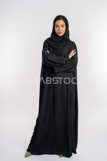 Expressions of self-confidence, modern and elegant veiled fashion, the concept of elegance and interest in external appearance, a portrait of a veiled Arab Gulf Emirati woman wearing an abaya and looking at something with gestures of firmness and seriousness, a young Saudi woman standing with crossed hands, a full body image, a white background