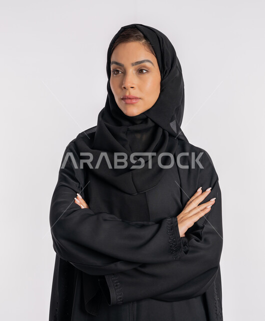 إيماءات الثقة بالنفس، أزياء محجبات عصرية راقية، مفهوم الأناقة والاهتمام بالمظهر الخارجي، بورتريه مقرب لامرأة عربية خليجية إماراتية محجبة ترتدي العباءة الحديثة وتنظر الى شيء ما بتعابير الحزم والجدية، شابة سعودية تقف بأيدي متقاطعة، خلفية بيضاء