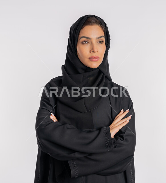 إيماءات الثقة بالنفس، أزياء محجبات عصرية راقية، مفهوم الأناقة والاهتمام بالمظهر الخارجي، بورتريه مقرب لامرأة عربية خليجية إماراتية محجبة ترتدي العباءة الحديثة وتنظر الى شيء ما بتعابير الحزم والجدية، شابة سعودية تقف بأيدي متقاطعة، خلفية بيضاء