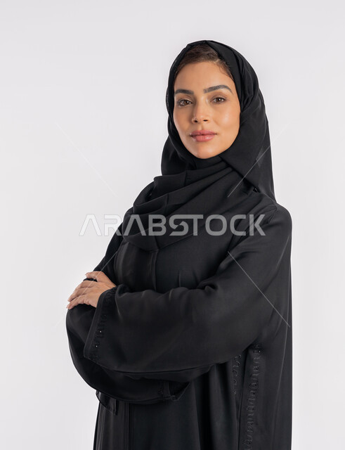 نظرات وتعابير السرور، أزياء محجبات عصرية راقية، مفهوم الأناقة والاهتمام بالمظهر الخارجي، بورتريه مقرب لامرأة عربية خليجية إماراتية مبتسمة ترتدي الحجاب والعباءة الحديثة وتنظر الى الكاميرا بإيماءات الثقة بالنفس، شابة سعودية تقف بأيدي متقاطعة، خلفية بيضاء