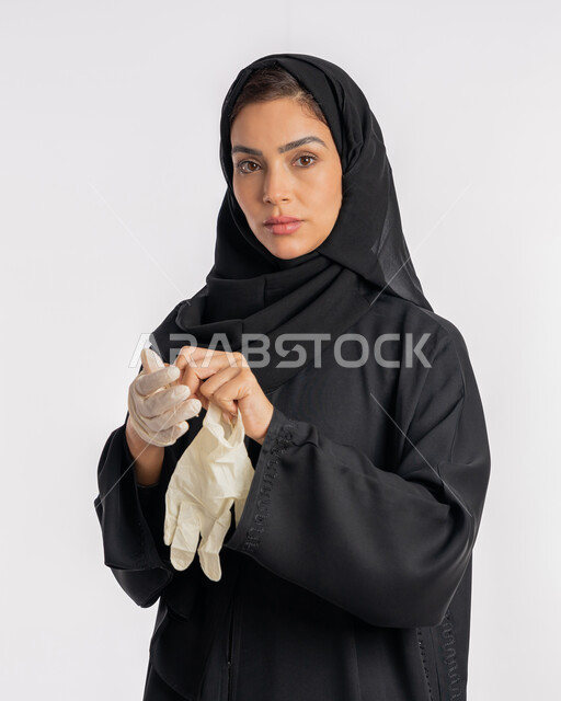 الحيطة من انتقال المرض, سيدة سعودية تتوخى الحذر من انتشار فيروس كورونا, بورتريه مقرب لإمرأة مقرب عربية خليجية إماراتية ترتدي الحجاب والعباءة تقوم بخلع القفازات وتبديلها, منع وتجنب ملامسة الايدي للجراثيم, اتباع إجراءات السلامة للوقاية من العدوى, خلفية بيضاء
