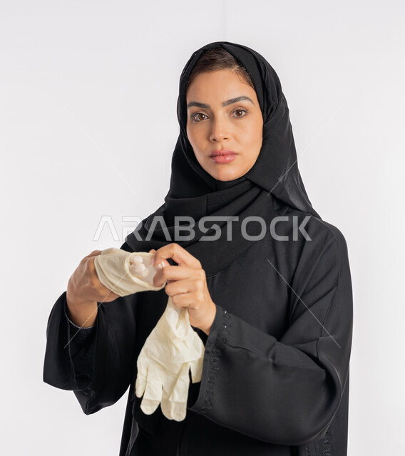 الحيطة من انتقال المرض, سيدة سعودية تتوخى الحذر من انتشار فيروس كورونا, بورتريه مقرب لإمرأة مقرب عربية خليجية إماراتية ترتدي الحجاب والعباءة تقوم بخلع القفازات وتبديلها, منع وتجنب ملامسة الايدي للجراثيم, اتباع إجراءات السلامة للوقاية من العدوى, خلفية بيضاء