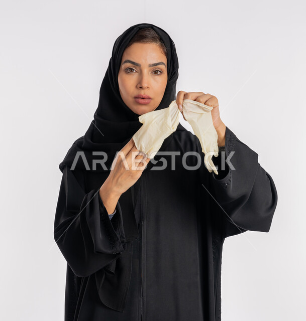 Caution against disease transmission, Saudi woman taking precautions against the spread of coronavirus, portrait of a close-up Arab Gulf Emirati woman wearing a hijab and abaya taking off and changing gloves, preventing and avoiding hands from touching germs, following safety measures to prevent infection, white background