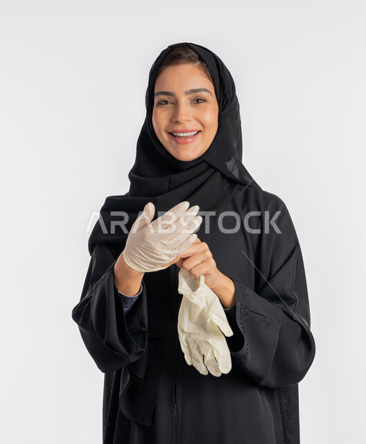 منع وتجنب ملامسة الايدي للجراثيم, اتباع إجراءات السلامة للوقاية من العدوى، الحيطة من انتقال المرض, سيدة سعودية تتوخى الحذر من انتشار فيروس كورونا, بورتريه مقرب لإمرأة مقرب عربية خليجية إماراتية ترتدي الحجاب والعباءة تقوم بخلع القفازات وتبديلها, خلفية بيضاء