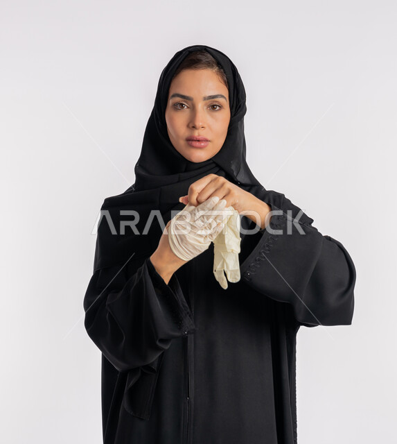منع وتجنب ملامسة الايدي للجراثيم, اتباع إجراءات السلامة للوقاية من العدوى، الحيطة من انتقال المرض, سيدة سعودية تتوخى الحذر من انتشار فيروس كورونا, بورتريه مقرب لإمرأة مقرب عربية خليجية إماراتية ترتدي الحجاب والعباءة تقوم بخلع القفازات وتبديلها, خلفية بيضاء