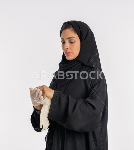 منع وتجنب ملامسة الايدي للجراثيم, اتباع إجراءات السلامة للوقاية من العدوى، الحيطة من انتقال المرض, سيدة سعودية تتوخى الحذر من انتشار فيروس كورونا, بورتريه مقرب لإمرأة مقرب عربية خليجية إماراتية ترتدي الحجاب والعباءة تقوم بخلع القفازات وتبديلها, خلفية بيضاء