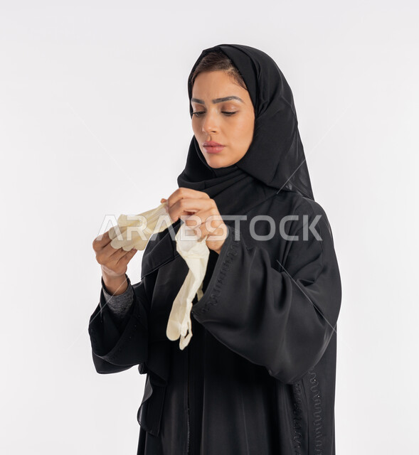 اتباع إجراءات السلامة للوقاية من العدوى، منع وتجنب ملامسة الايدي للجراثيم, الحيطة من انتقال المرض, سيدة سعودية تتوخى الحذر من انتشار فيروس كورونا, بورتريه لإمرأة مقرب عربية خليجية إماراتية ترتدي الحجاب والعباءة تقوم بخلع القفازات وتبديلها, خلفية بيضاء