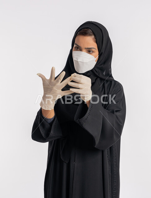 سيدة سعودية تتوخى الحذر من انتشار فيروس كورونا, بورتريه مقرب لإمرأة مقرب عربية خليجية إماراتية ترتدي الحجاب والعباءة والكمامة تقوم بارتداء القفازات, منع وتجنب ملامسة الايدي للجراثيم, اتباع إجراءات السلامة للوقاية من العدوى, خلفية بيضاء