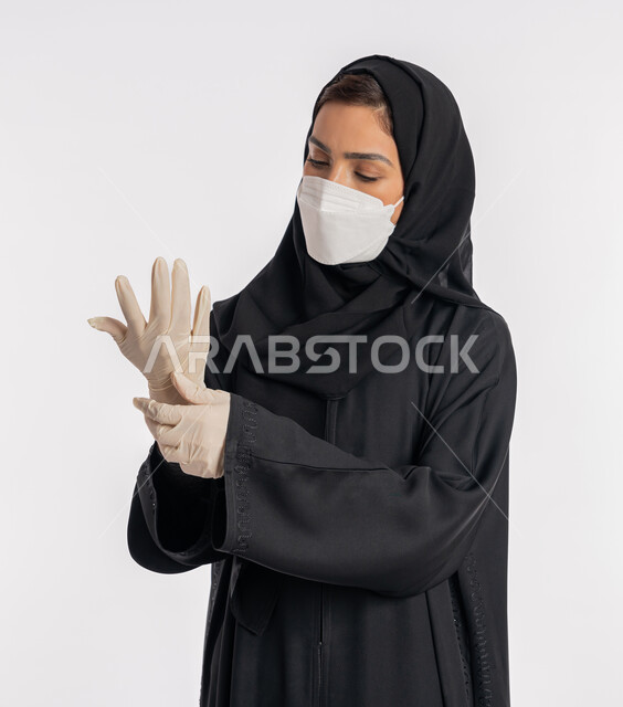 سيدة سعودية تتوخى الحذر من انتشار فيروس كورونا, بورتريه مقرب لإمرأة مقرب عربية خليجية إماراتية ترتدي الحجاب والعباءة والكمامة تقوم بارتداء القفازات, منع وتجنب ملامسة الايدي للجراثيم, اتباع إجراءات السلامة للوقاية من العدوى, خلفية بيضاء