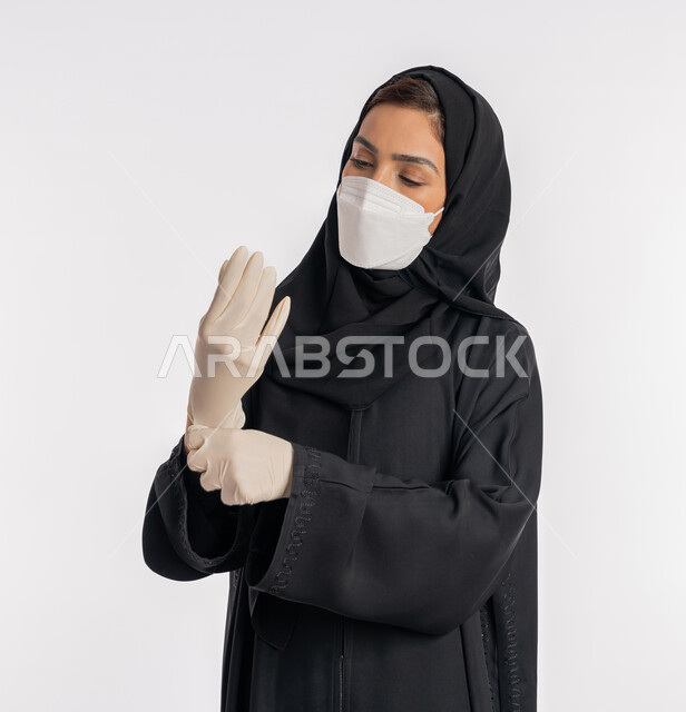 Saudi woman taking precautions against the spread of coronavirus, portrait of a close-up Arab Gulf Emirati woman wearing a hijab, abaya and mask wearing gloves, preventing and avoiding hands from touching germs, following safety measures to prevent infection, white background