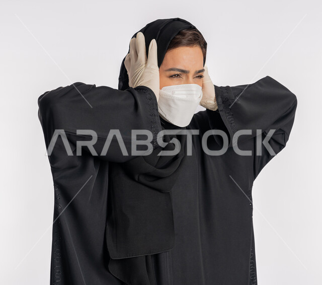 تعابير الاستياء والضجر من المرض، شابة سعودية تتبع الاجراءات الوقائية من فايروس كورونا، الصداع وألم الرأس والمعاناة من آلام الشقيقة، بورتريه مقرب لإمرأة عربية خليجية إماراتية محجبة ترتدي العباءة والكمامة تضع يديها على رأسها بايماءات الارهاق، خلفية بيضاء