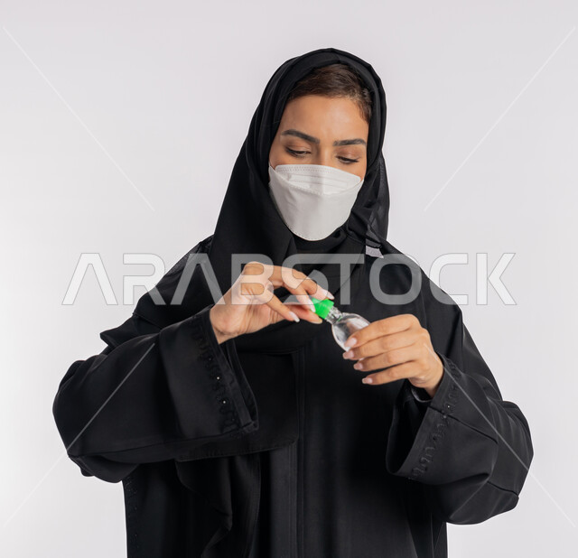 استخدام المواد الكيميائية والكحول للقضاء على الفايروسات والجراثيم، شابة سعودية تتبع الاجراءات الاحترازية للوقاية من فايروس كورونا، بورتريه مقرب لإمرأة عربية خليجية إماراتية ترتدي العباءة والحجاب تضع الكمامة تمسك علبة المعقم بيدها, خلفية بيضاء