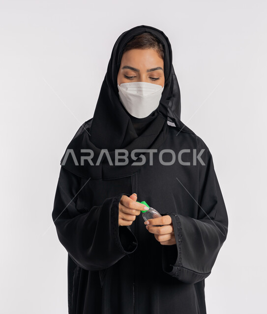 استخدام المواد الكيميائية والكحول للقضاء على الفايروسات والجراثيم، شابة سعودية تتبع الاجراءات الاحترازية للوقاية من فايروس كورونا، بورتريه مقرب لإمرأة عربية خليجية إماراتية ترتدي العباءة والحجاب تضع الكمامة تمسك علبة المعقم بيدها, خلفية بيضاء