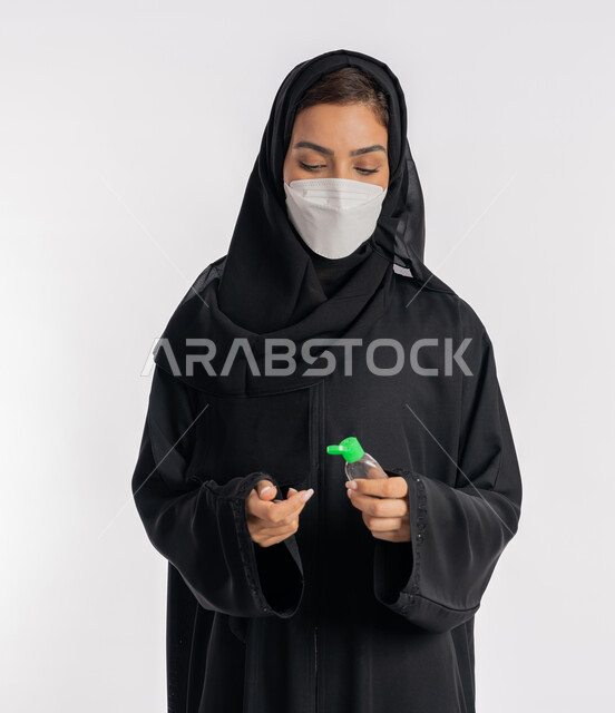 استخدام المواد الكيميائية والكحول للقضاء على الفايروسات والجراثيم، شابة سعودية تتبع الاجراءات الاحترازية للوقاية من فايروس كورونا، بورتريه مقرب لإمرأة عربية خليجية إماراتية ترتدي العباءة والحجاب تضع الكمامة تمسك علبة المعقم بيدها, خلفية بيضاء